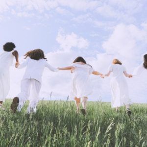 Grupo de mujeres de diversas etnias tomadas de la mano, vestidas de blanco, corriendo por un campo verde bajo un cielo despejado.